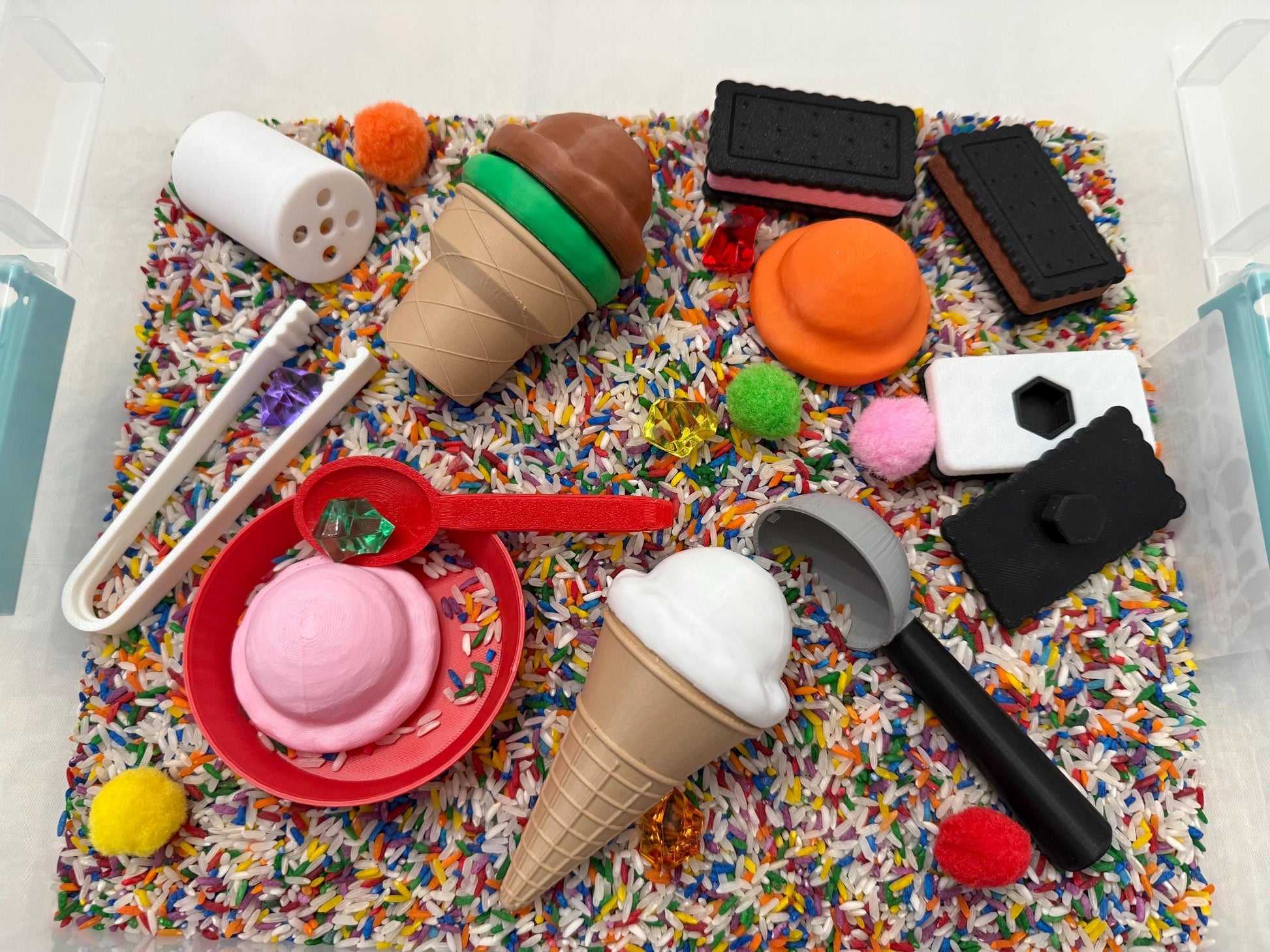 May include: A sensory bin filled with colorful rice and sprinkles, featuring various ice cream cones, scoops, and other small toys.