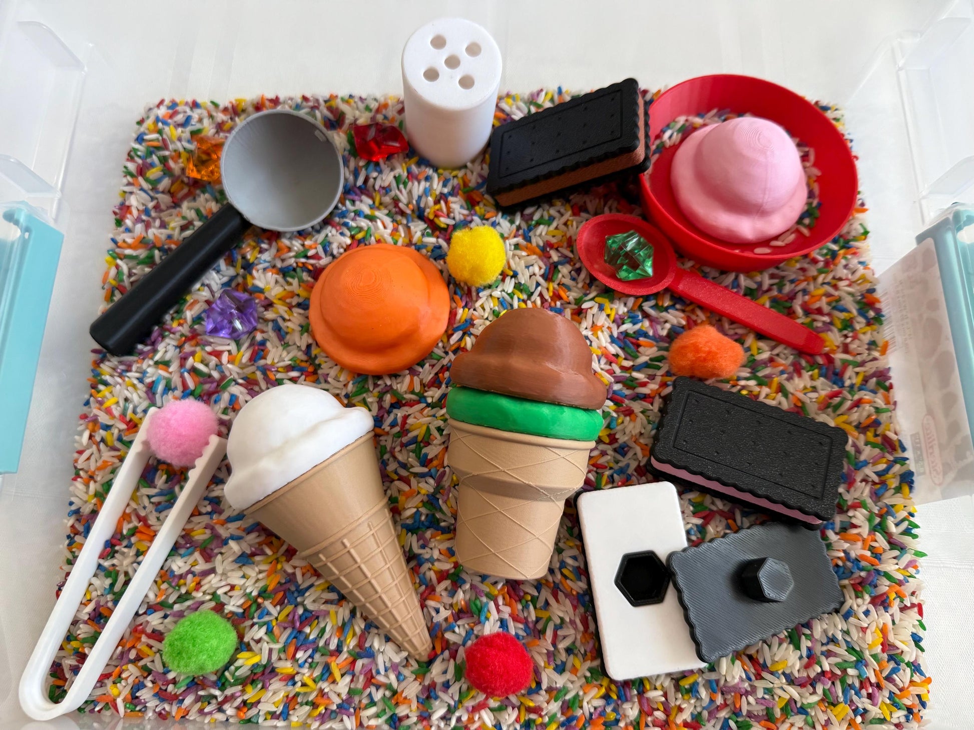 May include: A sensory bin filled with colorful sprinkles and small toys, including two ice cream cones, a scoop, a small container, and a few chocolate bars.