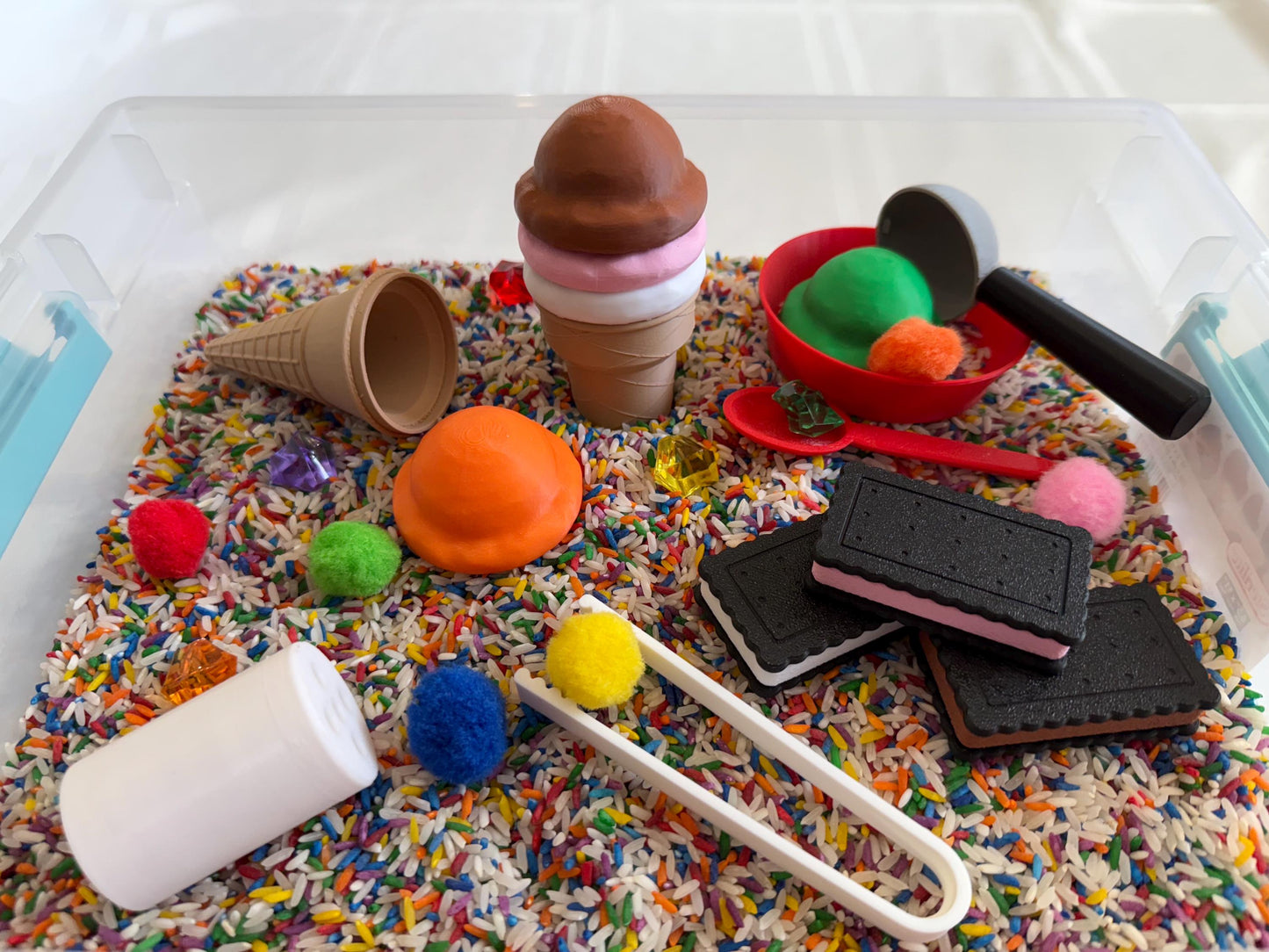 May include: A sensory bin filled with colorful rice and sprinkles, featuring a variety of toys including an ice cream cone, a scoop, a small bowl, and a few cookies. There are also several pom-poms and a pair of tongs.