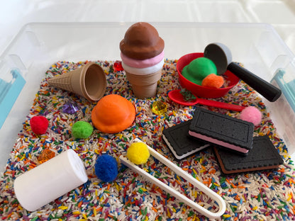 May include: A sensory bin filled with colorful rice and sprinkles, featuring a variety of toys including an ice cream cone, a scoop, a small bowl, and a few cookies. There are also several pom-poms and a pair of tongs.