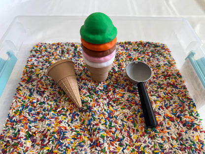 May include: A plastic ice cream cone with four scoops of play dough in green, orange, brown, and pink colors. The ice cream cone is sitting in a clear plastic container filled with colorful rice. A brown plastic ice cream cone and a gray plastic ice cream scoop are also in the container.