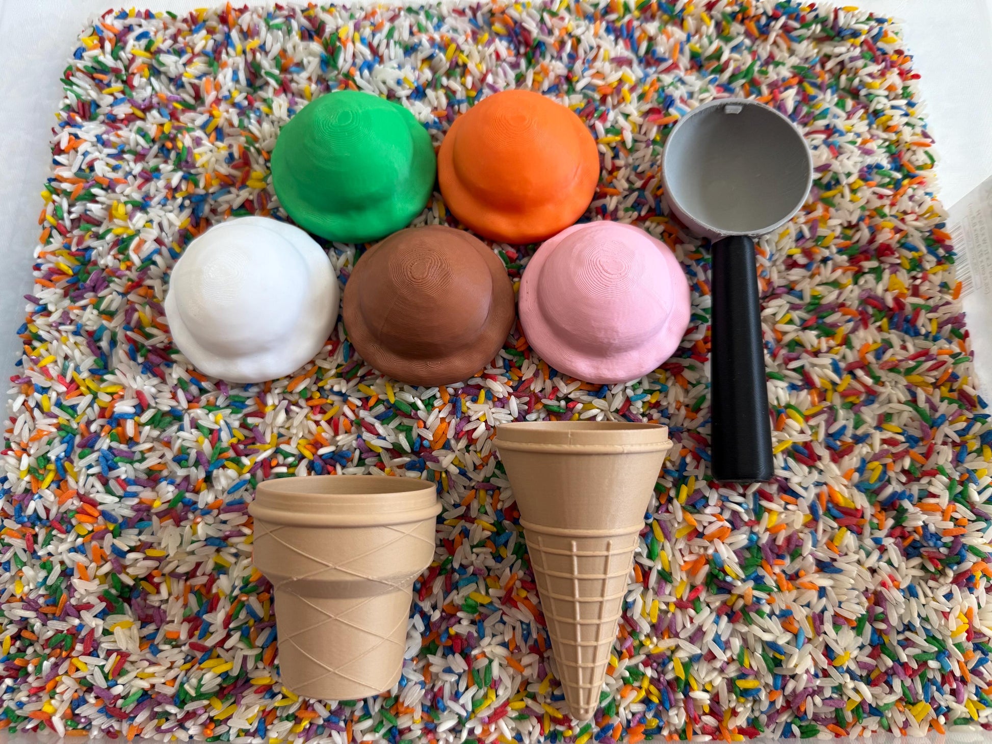 May include: A set of six colorful ice cream cones and a scoop, all made of plastic, on a bed of colorful sprinkles. The cones are in various colors, including green, orange, brown, white, and pink.