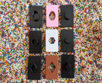 May include: Nine different shaped cookie cutters, each with a different design. The cutters are arranged in three rows of three. The cutters are made of black, white, pink, and brown plastic. The cutters are on a background of colorful rice.