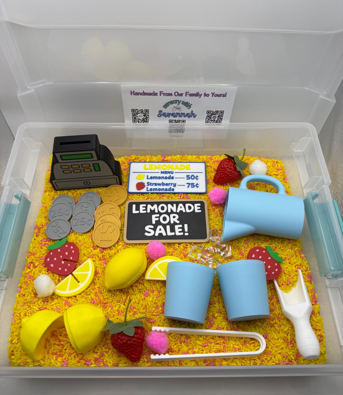May include: A sensory bin with a lemonade stand theme. Includes a toy cash register, coins, lemons, strawberries, a pitcher, cups, and a sign that reads "Lemonade for Sale!". The bin is filled with yellow rice.