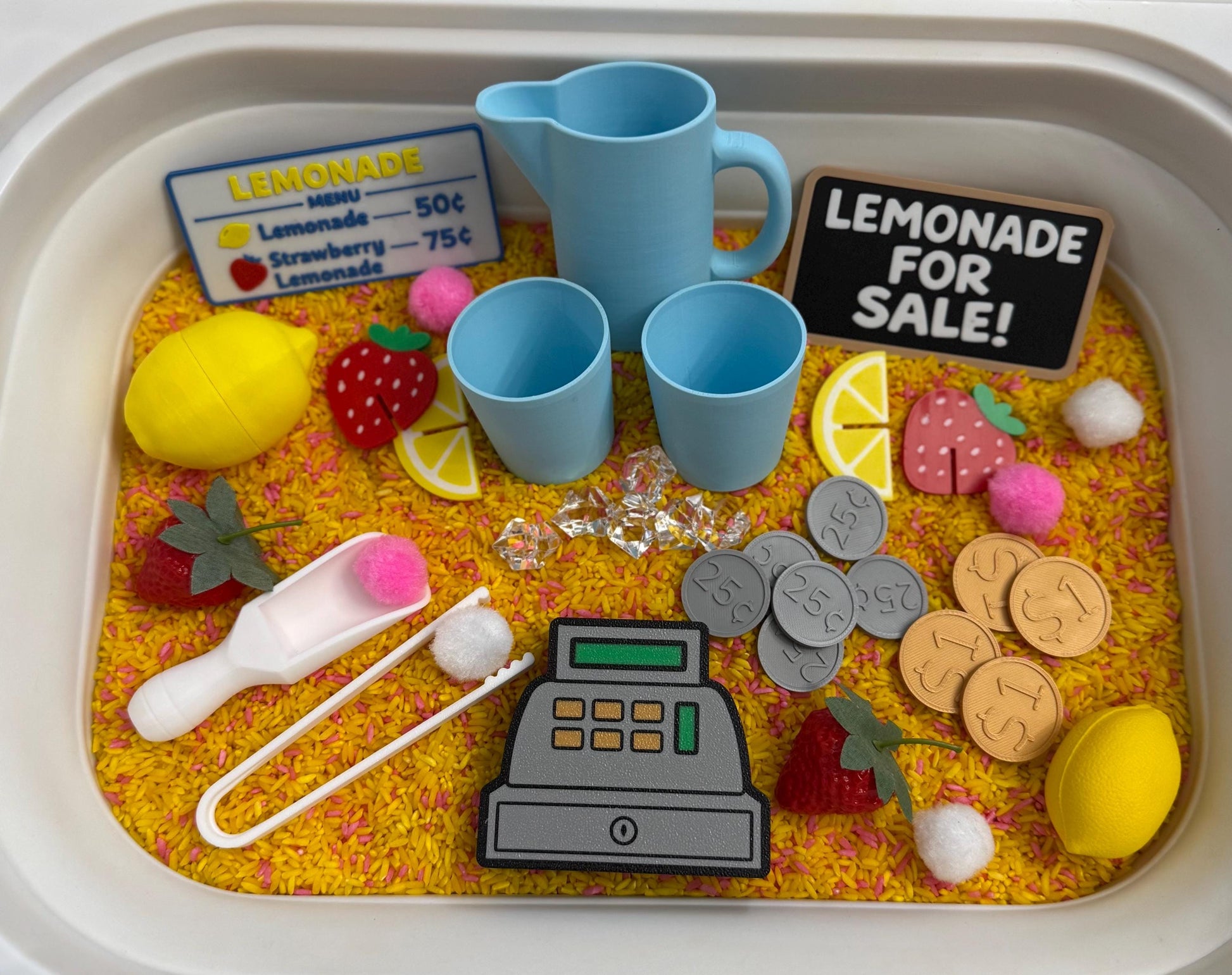 May include: A play lemonade stand set with a blue pitcher, cups, and a sign that says "Lemonade For Sale!". Includes toy lemons, strawberries, coins, and a cash register. The set is arranged on a bed of yellow and orange rice.