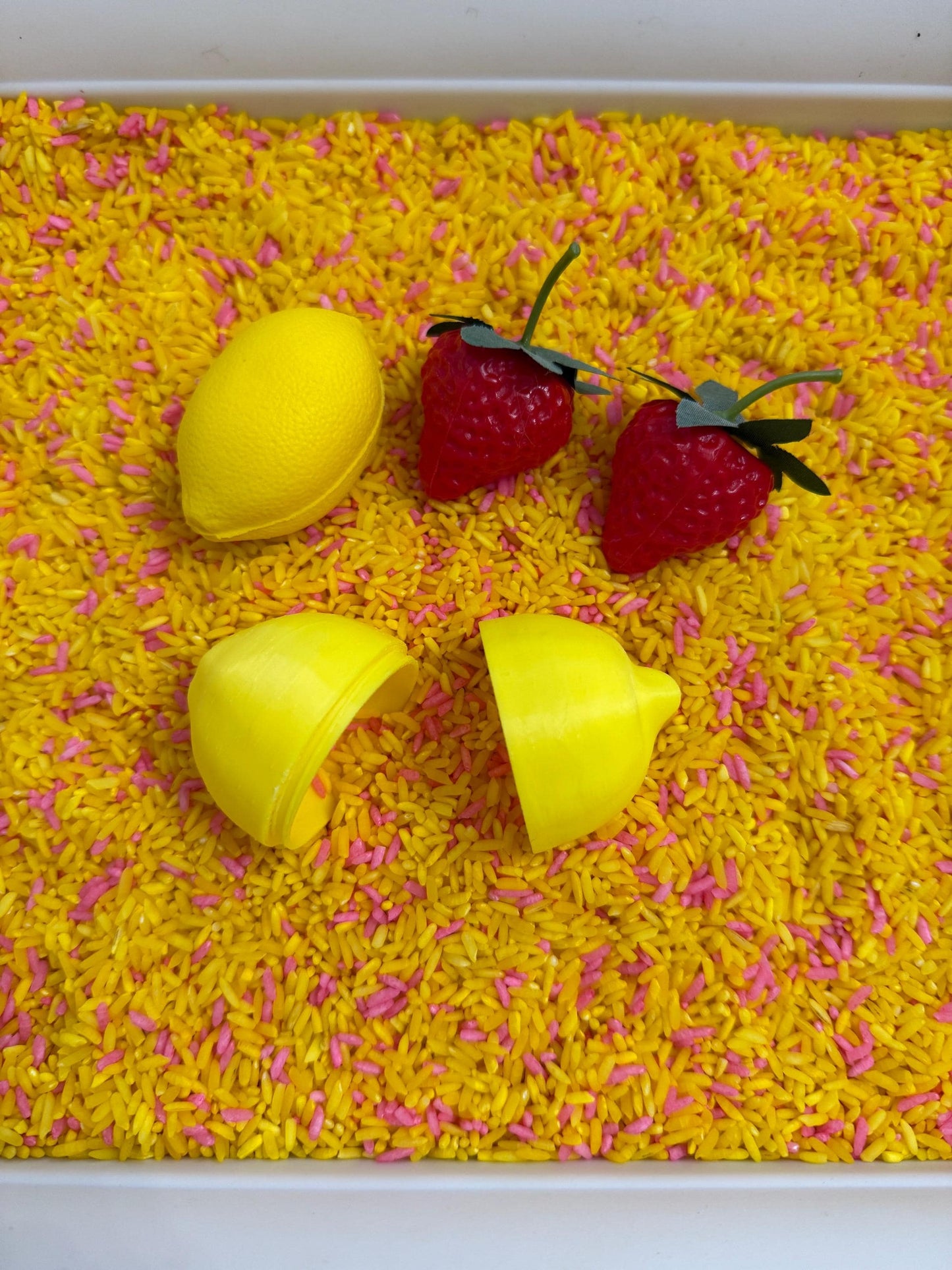May include: A sensory bin filled with yellow and pink dyed rice. The bin contains a yellow lemon, two red strawberries, and a yellow lemon cut in half. The items are arranged on the rice.