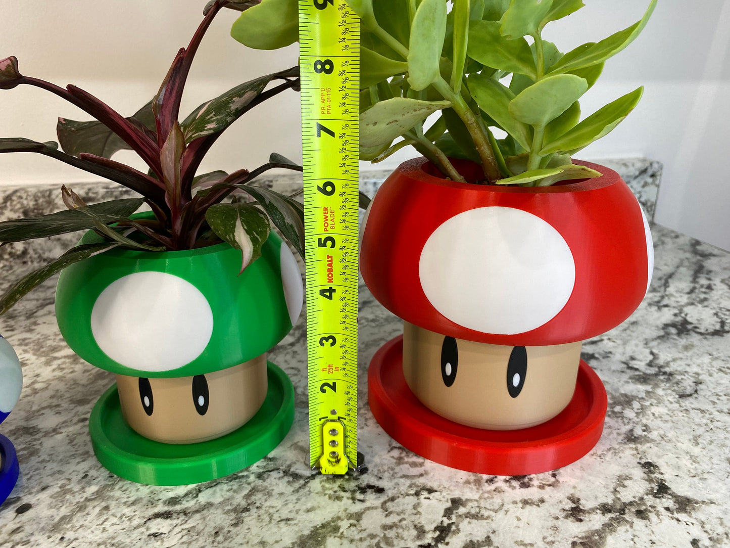 May include: Two mushroom-shaped plant pots, one green and one red, with white spots and brown faces. The pots are 5 inches tall and are sitting on a tiled surface.