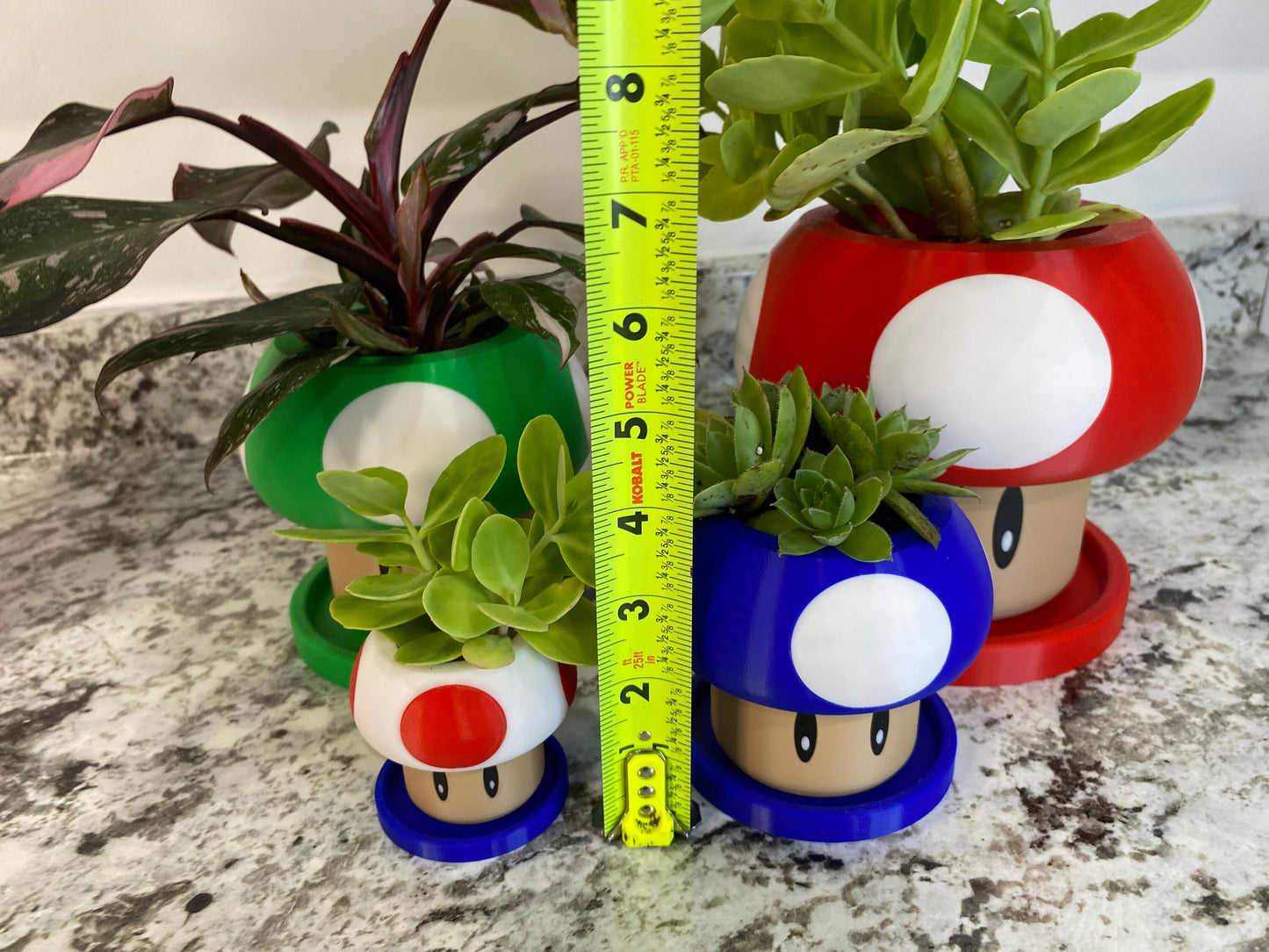May include: Three different sized mushroom-shaped planters, each with a different colored top. The largest planter is red with a white dot, the middle planter is green with a white dot, and the smallest planter is blue with a white dot. All three planters have a brown base.