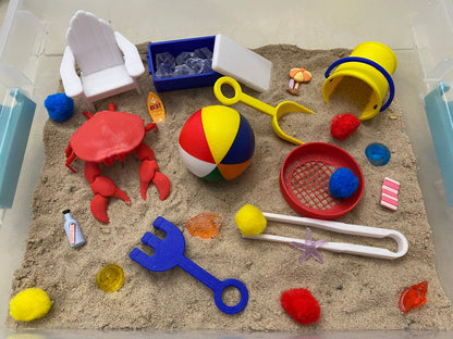 May include: A beach-themed sensory bin with a red crab, a blue and yellow beach ball, a blue and yellow pail, a blue and yellow shovel, a red and white striped beach towel, a white beach chair, a blue and white cooler, a white pair of tongs, a red strainer, a blue and white surfboard, a starfish, and a variety of pom-poms.