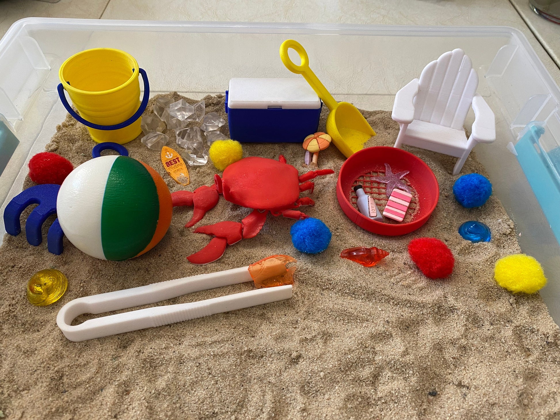 May include: A beach-themed sensory bin with a red crab, a beach ball, a yellow bucket, a blue cooler, a white beach chair, a yellow shovel, a red strainer, a pair of tweezers, and various colorful pom-poms.