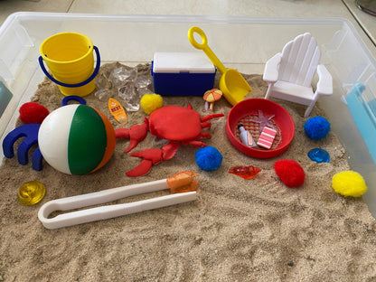May include: A beach-themed sensory bin with a red crab, a beach ball, a yellow bucket, a blue cooler, a white beach chair, a yellow shovel, a red strainer, a pair of tweezers, and various colorful pom-poms.