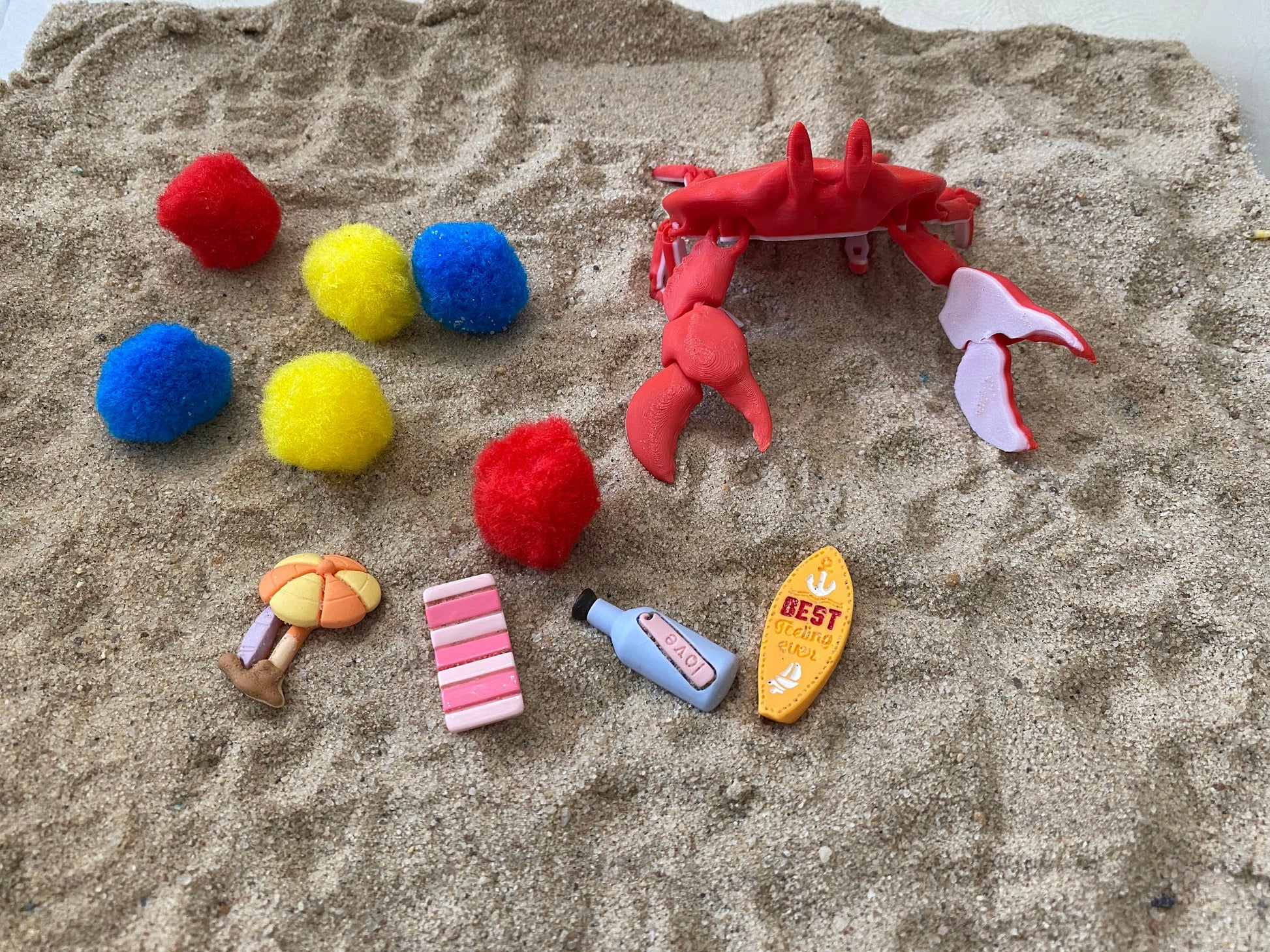 May include: A red plastic crab toy, four yellow pom-poms, two blue pom-poms, two red pom-poms, a pink and white striped beach towel, a yellow and orange beach umbrella, a blue bottle with a white label that says "BAOI", and a yellow surfboard with the text "BEST Surfing Ever!"