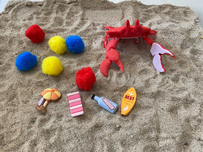 May include: A red plastic crab toy, four yellow pom-poms, two blue pom-poms, two red pom-poms, a pink and white striped beach towel, a yellow and orange beach umbrella, a blue bottle with a white label that says "BAOI", and a yellow surfboard with the text "BEST Surfing Ever!"
