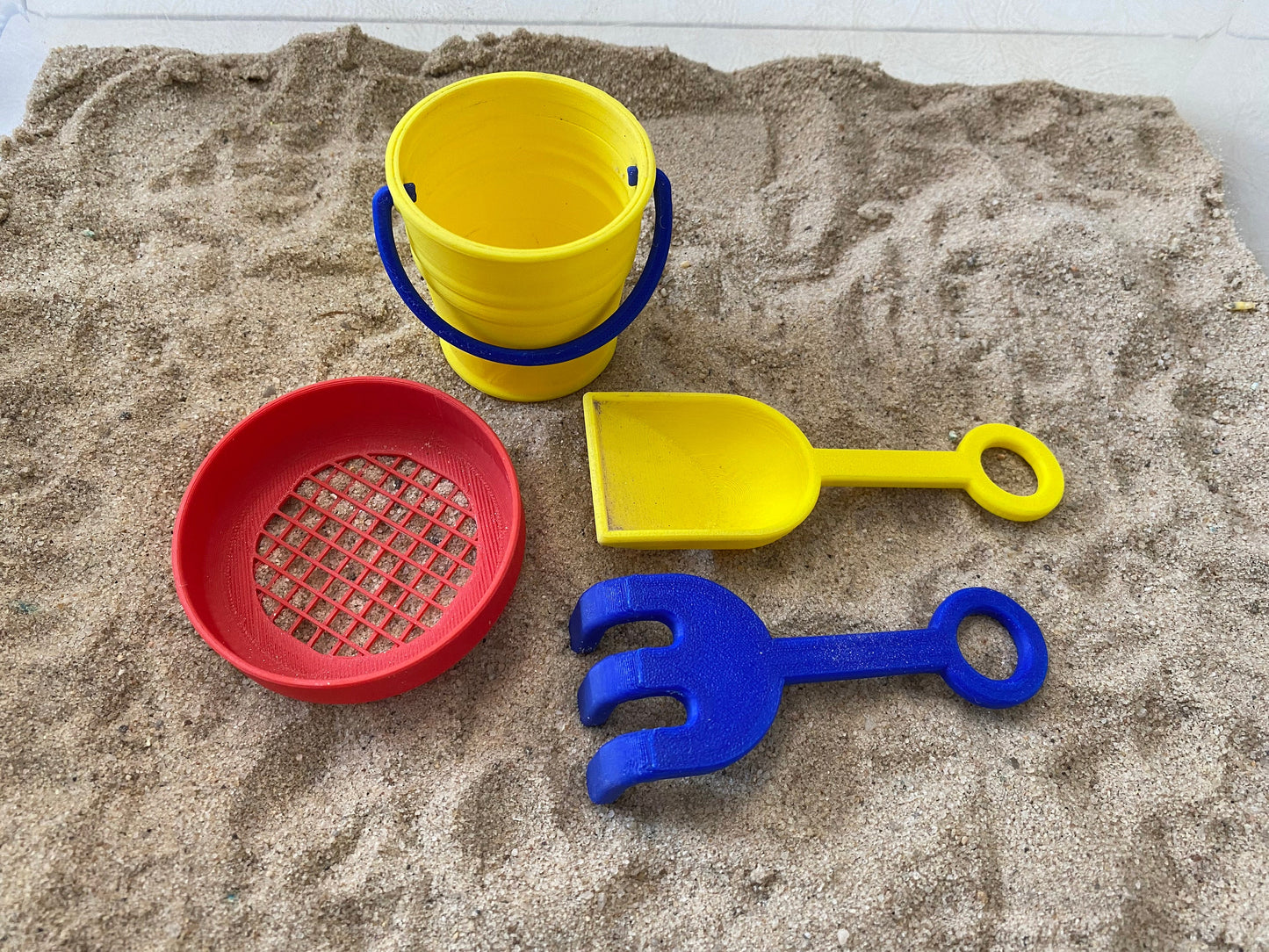 May include: A set of yellow and blue plastic beach toys, including a bucket, a shovel, a rake, and a sifter. The toys are on a bed of sand.