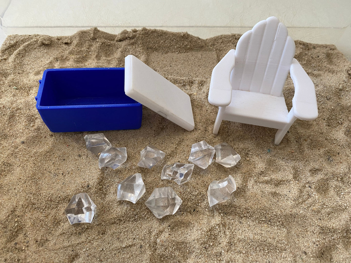 May include: A blue cooler with a white lid, a white plastic beach chair, and clear ice cubes on a sandy surface.