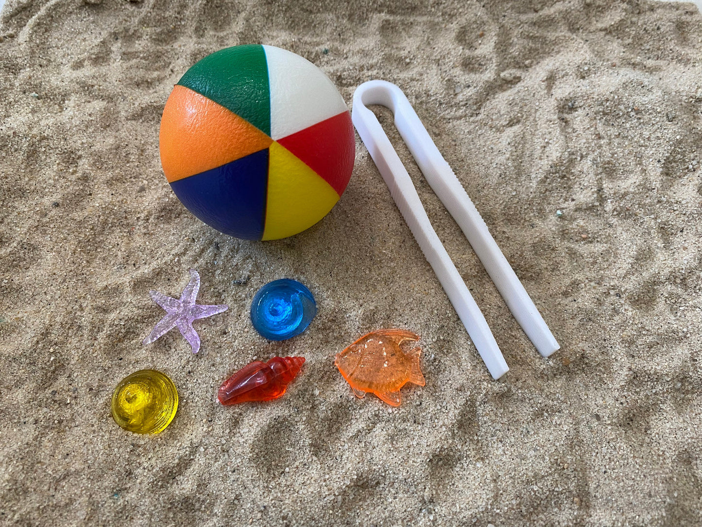 May include: A multi-colored beach ball with a white plastic pair of tweezers and a variety of small plastic sea life toys on a sandy surface.