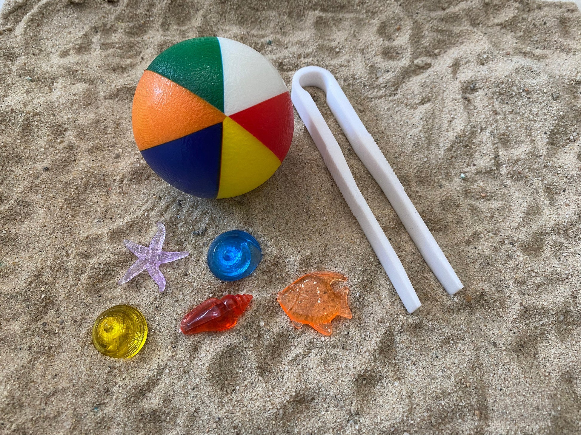 May include: A multi-colored beach ball with a white plastic pair of tweezers and a variety of small plastic sea life toys on a sandy surface.