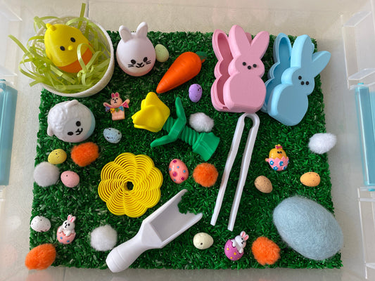 May include: A green sensory bin filled with rice, featuring Easter-themed toys, including a chick, bunnies, eggs, a carrot, a flower, and a scoop.