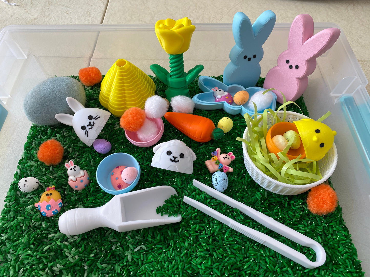 May include: A sensory bin filled with green rice, featuring a variety of Easter-themed toys, including bunnies, chicks, eggs, and a carrot. There are also colorful pom-poms, a scoop, and tongs for play.