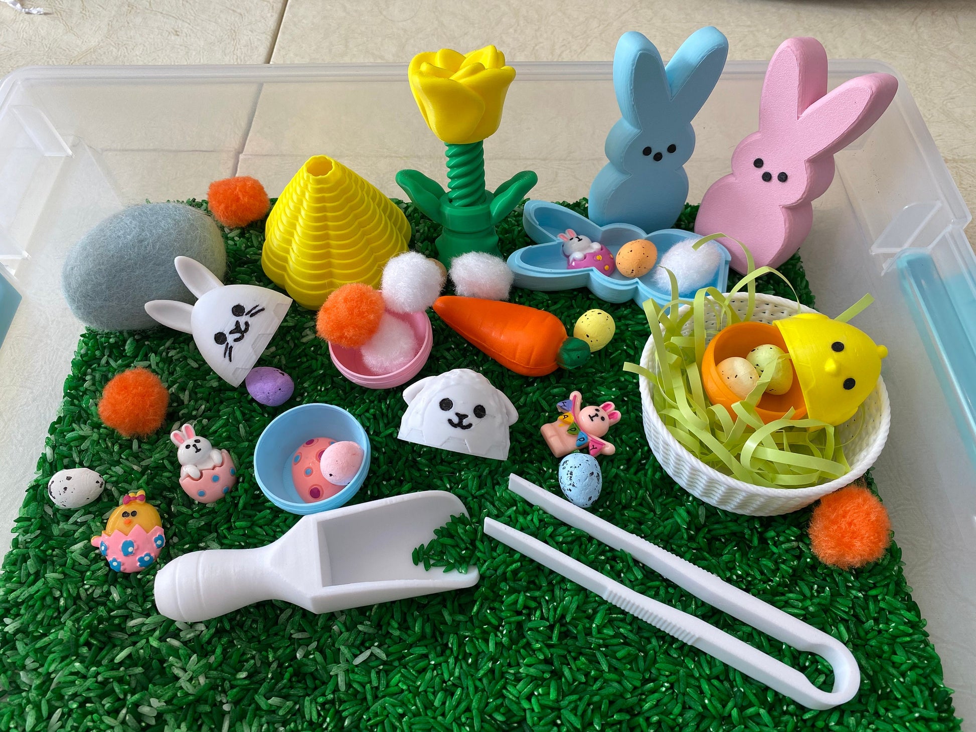 May include: A sensory bin filled with green rice, featuring a variety of Easter-themed toys, including bunnies, chicks, eggs, and a carrot. There are also colorful pom-poms, a scoop, and tongs for play.