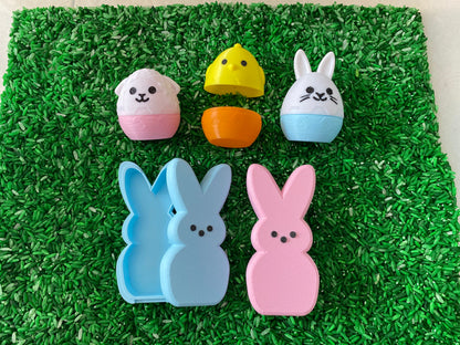 May include: A set of Easter-themed nesting eggs and bunny-shaped molds. The eggs are decorated with a chick, a lamb, and a bunny. The molds are blue, pink, and white.