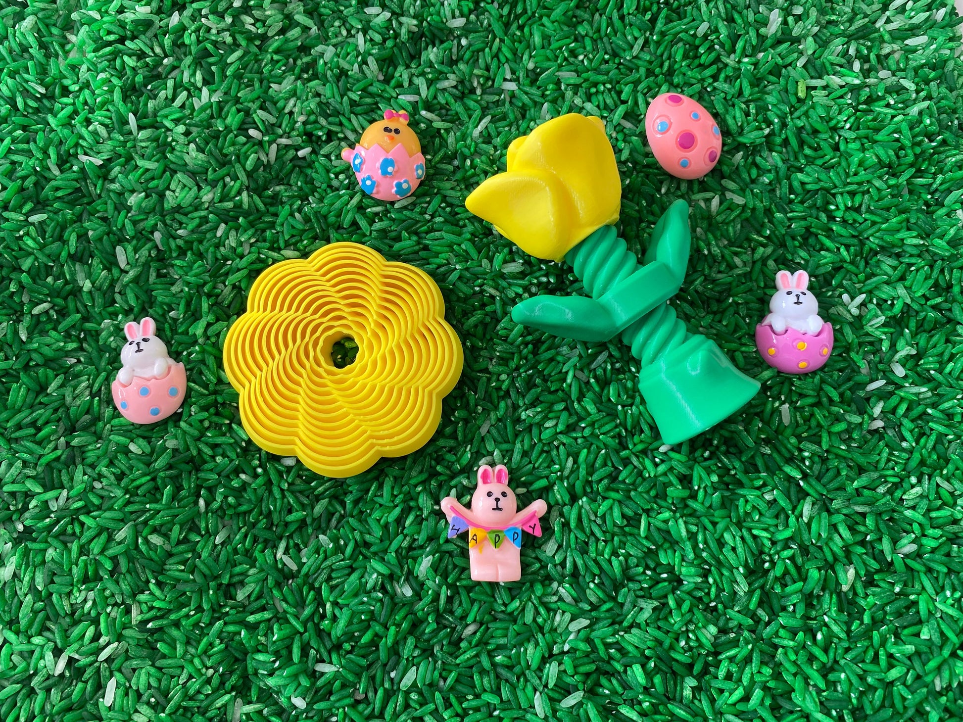 May include: A collection of Easter-themed toys on a green background. The toys include a yellow flower, a yellow tulip, a pink bunny holding a banner that says "Happy", a pink bunny in an egg, a yellow chick in an egg, and a pink egg with white polka dots.
