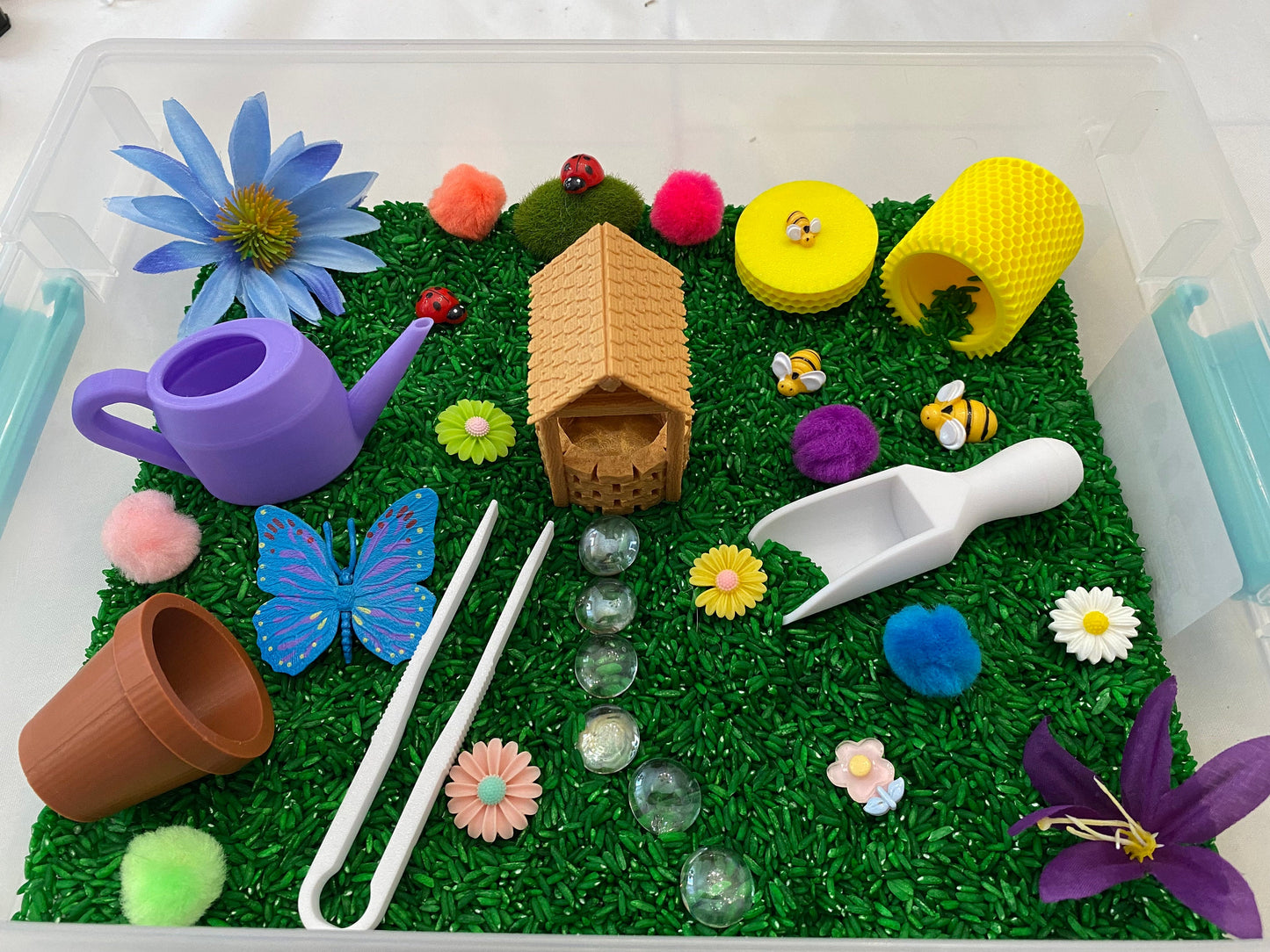 May include: A sensory bin filled with green rice, featuring a variety of miniature toys, including a house, a butterfly, a watering can, a bee, a ladybug, flowers, and a scoop.
