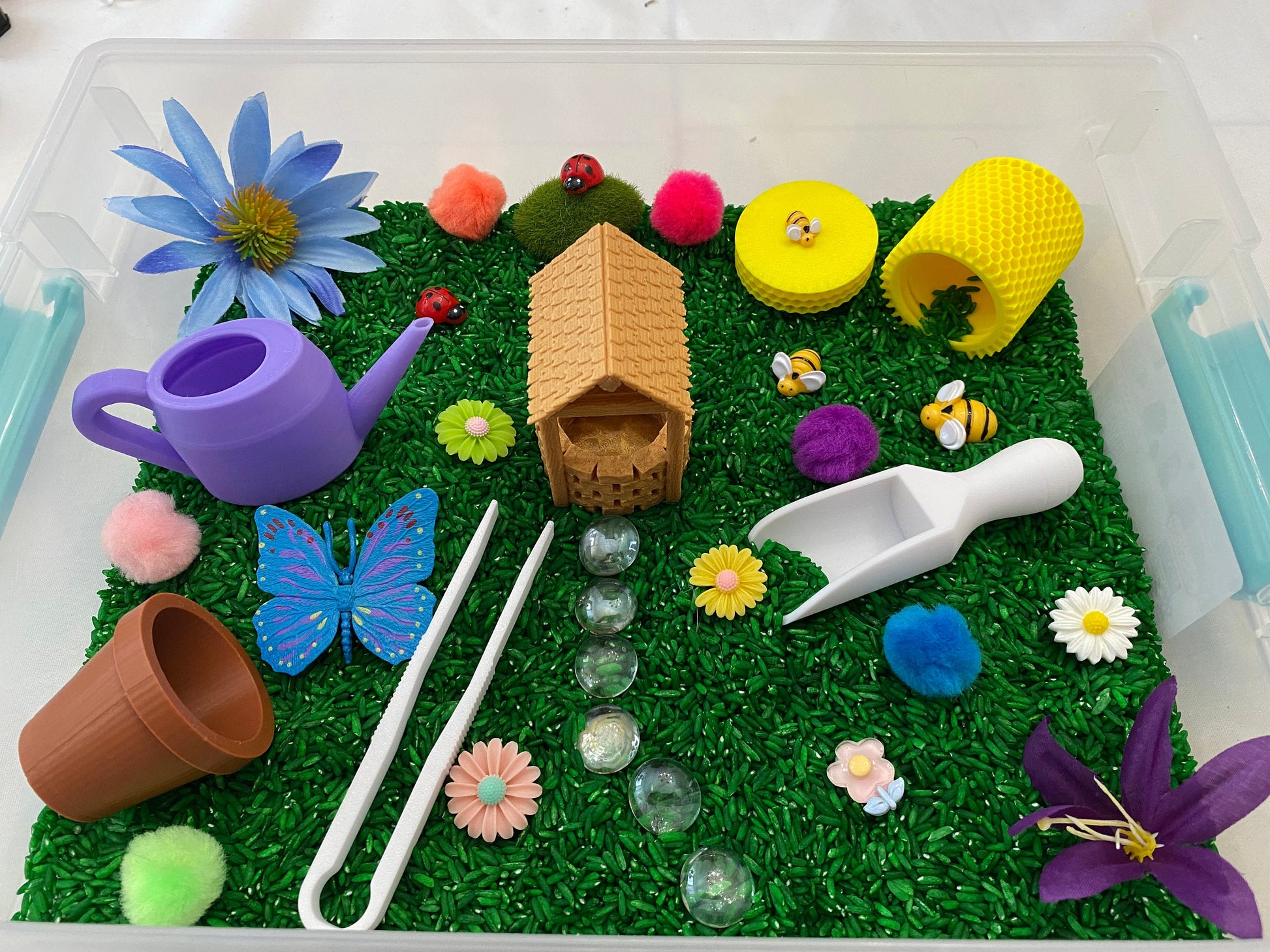 May include: A sensory bin filled with green rice, featuring a variety of miniature toys, including a house, a butterfly, a watering can, a bee, a ladybug, flowers, and a scoop.