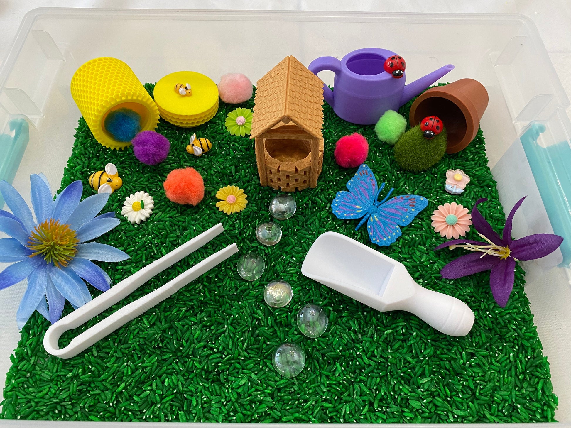 May include: A sensory bin filled with green rice, featuring a small wooden house, a purple watering can, a brown pot, a blue butterfly, a white scoop, white tongs, and various colorful flowers and pom-poms.
