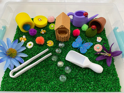May include: A sensory bin filled with green rice, featuring a small wooden house, a purple watering can, a brown pot, a blue butterfly, a white scoop, white tongs, and various colorful flowers and pom-poms.