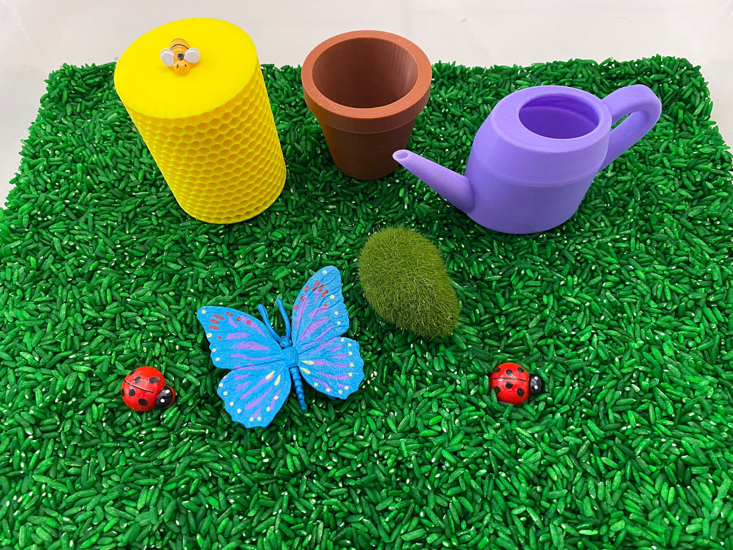 May include: A green sensory bin filled with green rice, a purple watering can, a brown flower pot, a yellow honeycomb container with a bee on top, a blue butterfly, and two red ladybugs.