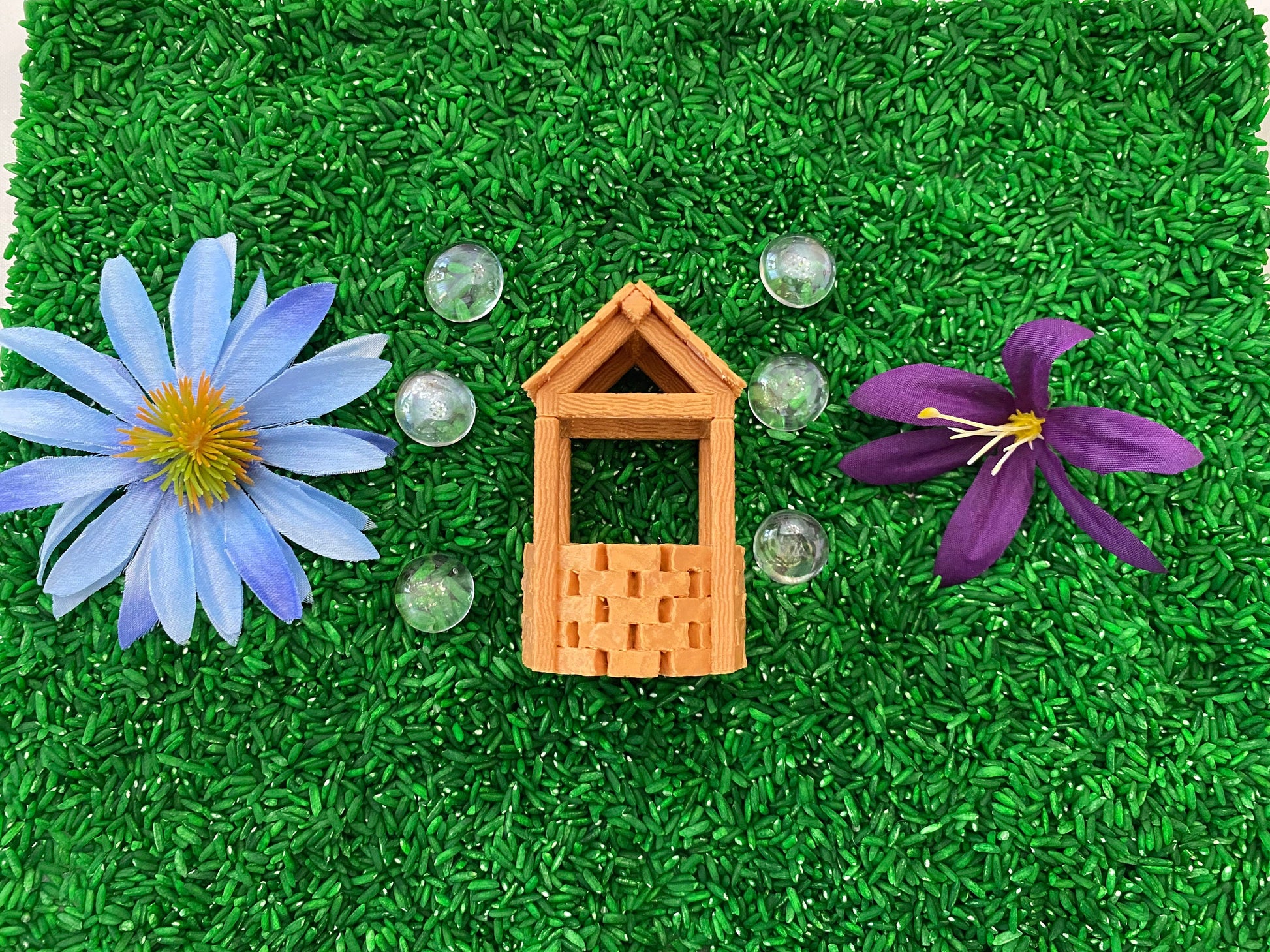 May include: A miniature wooden well with a brown roof, surrounded by green grass and blue and purple flowers. There are also several clear glass marbles scattered around the well.