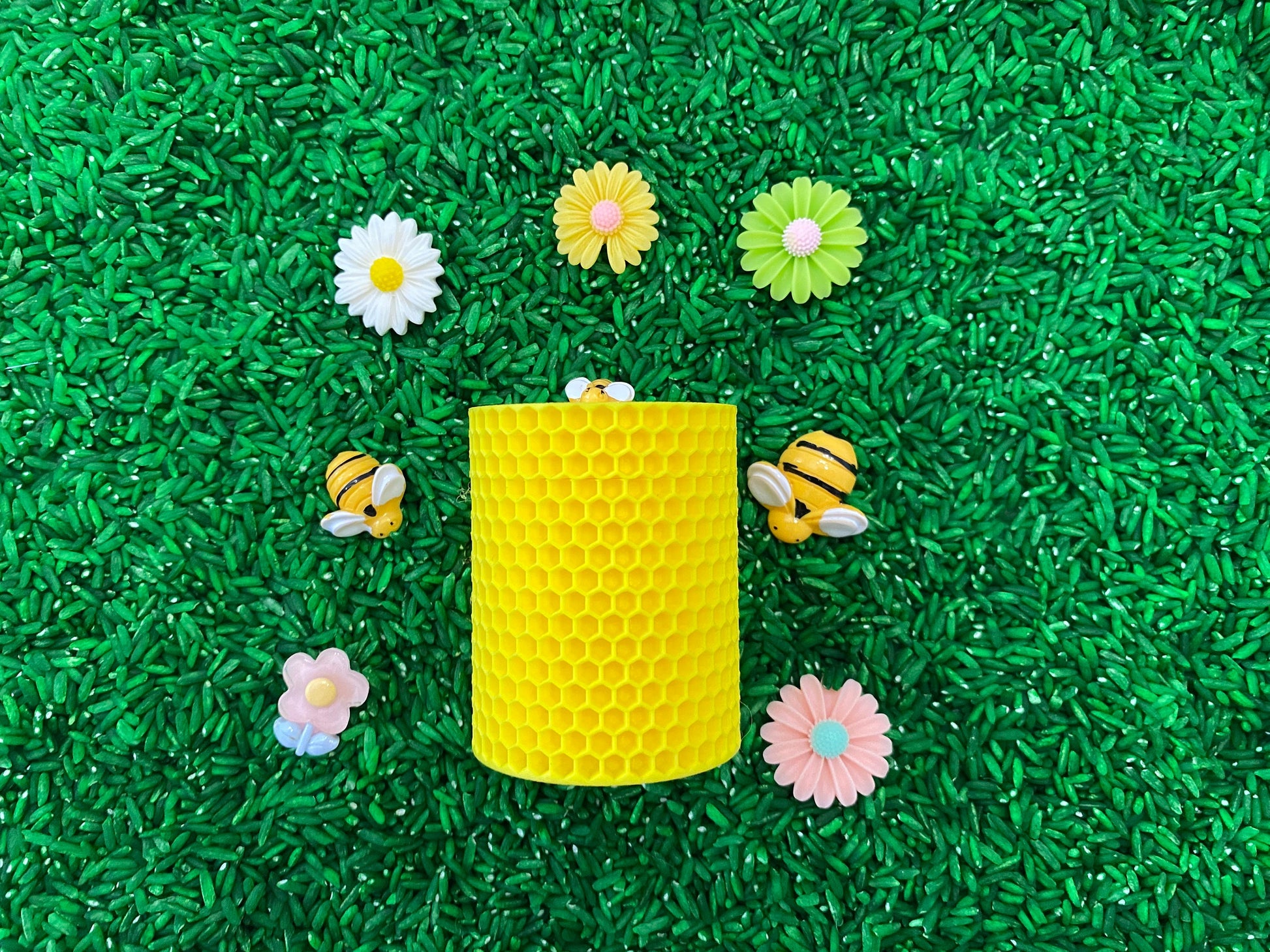 May include: A yellow honeycomb-shaped container surrounded by plastic bees and flowers on a green surface.