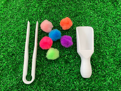 May include: A set of sensory tools for kids, including a white scoop, tweezers, and six colorful pom-poms in pink, orange, blue, purple, green, and light pink. The tools are on a green surface.