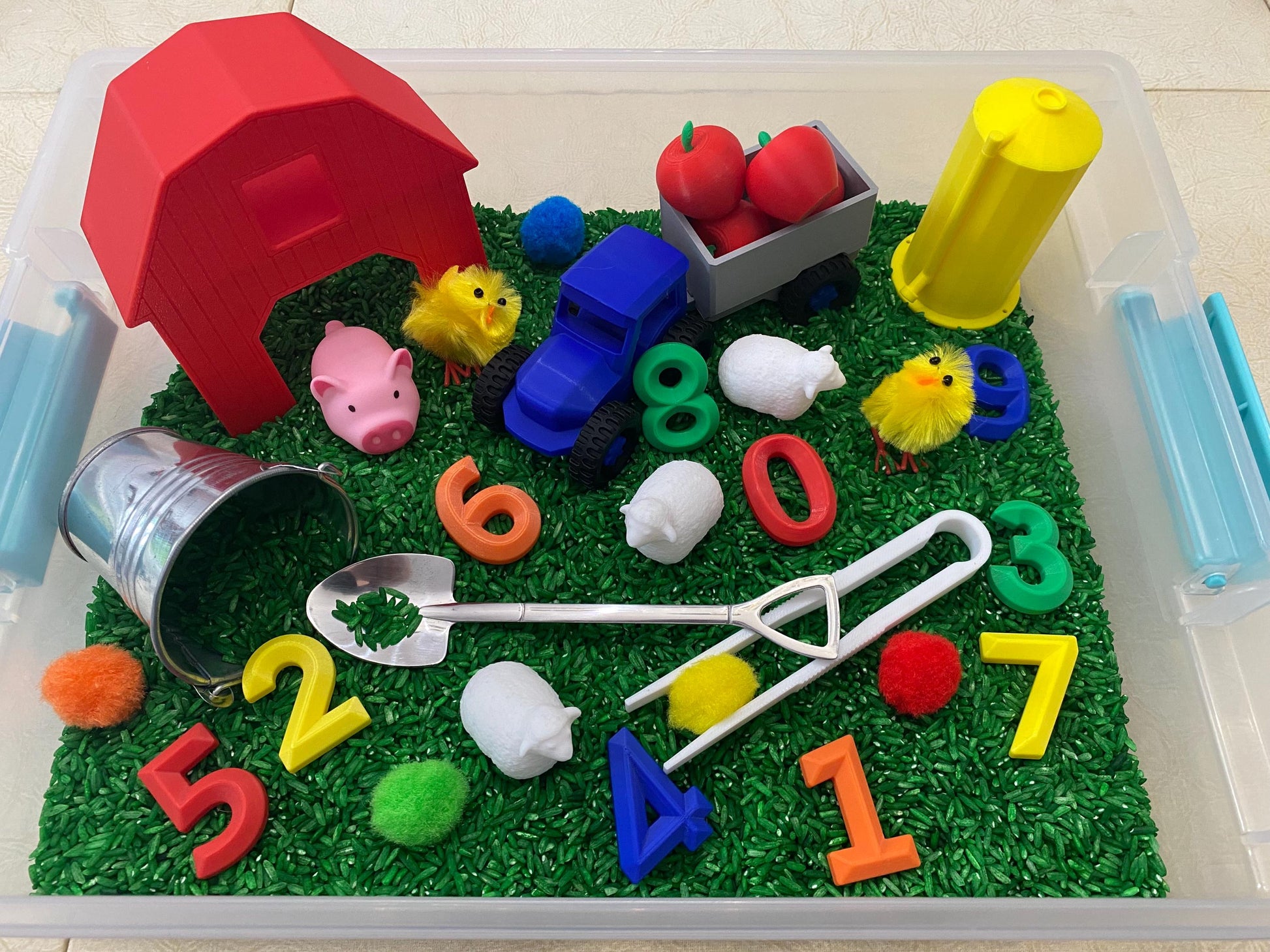 May include: A red barn, a blue toy truck, a bucket, a shovel, and a variety of farm animals are placed in a clear plastic bin filled with green rice. The numbers 1 through 8 are scattered throughout the bin.