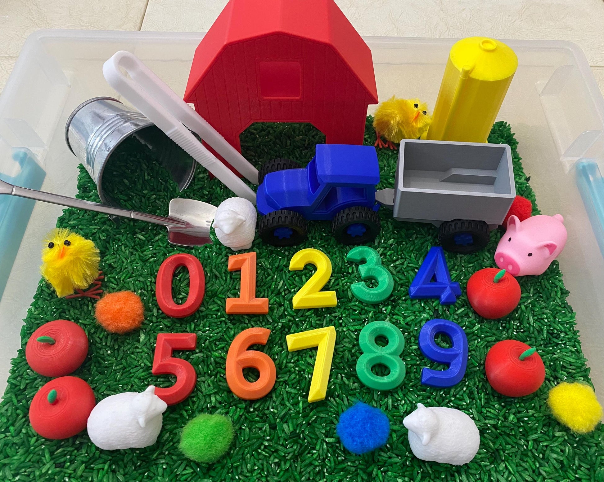 May include: A red barn, a blue tractor with a gray wagon, a yellow silo, and a set of colorful numbers 0-9 are in a sensory bin filled with green rice. There are also red apples, white lambs, and yellow chicks.