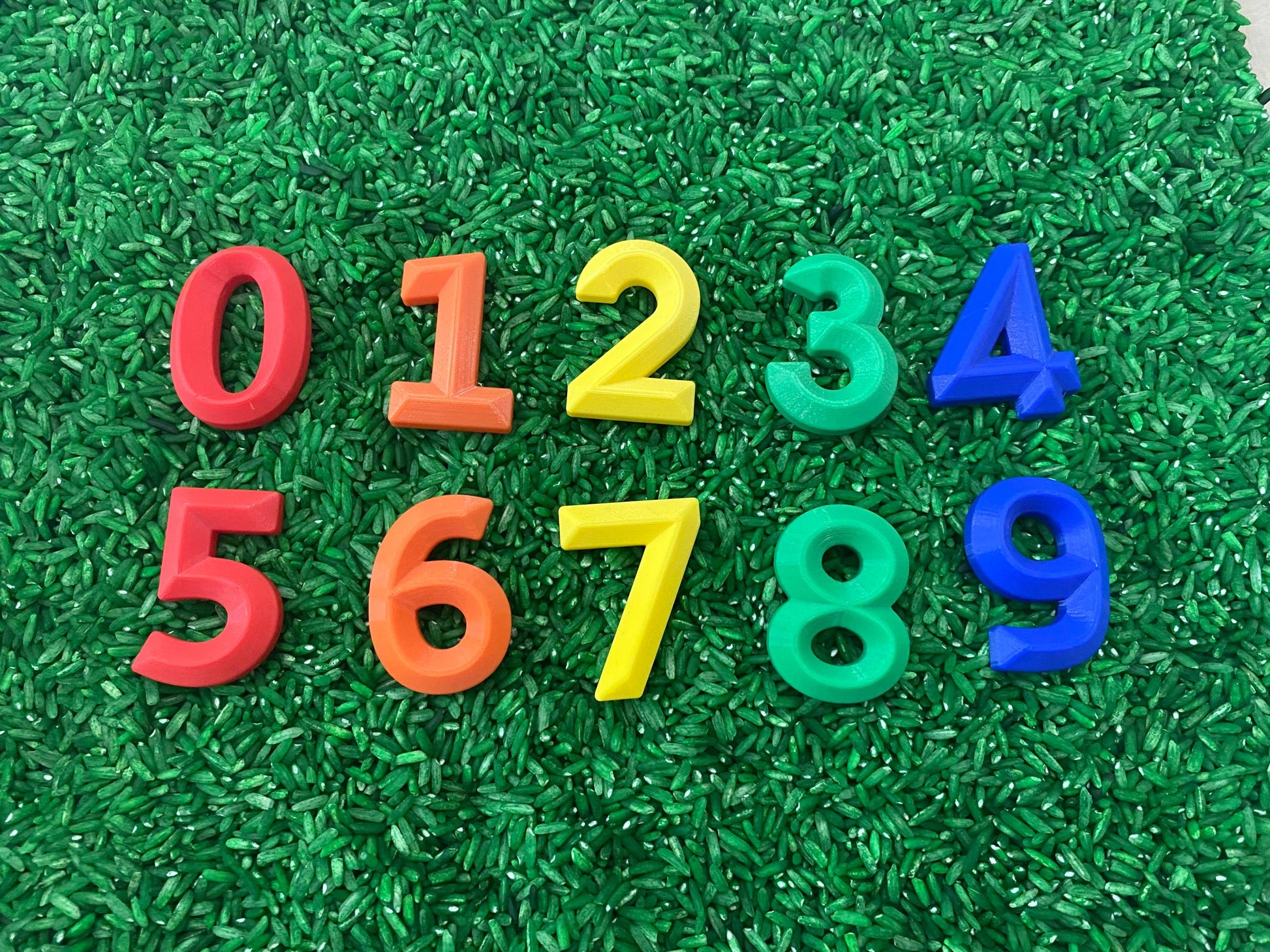 May include: Numbers 0 through 9 are displayed on a green surface. The numbers are made of plastic and are in different colors: red, orange, yellow, green, and blue.