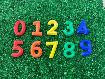 May include: Numbers 0 through 9 are displayed on a green surface. The numbers are made of plastic and are in different colors: red, orange, yellow, green, and blue.