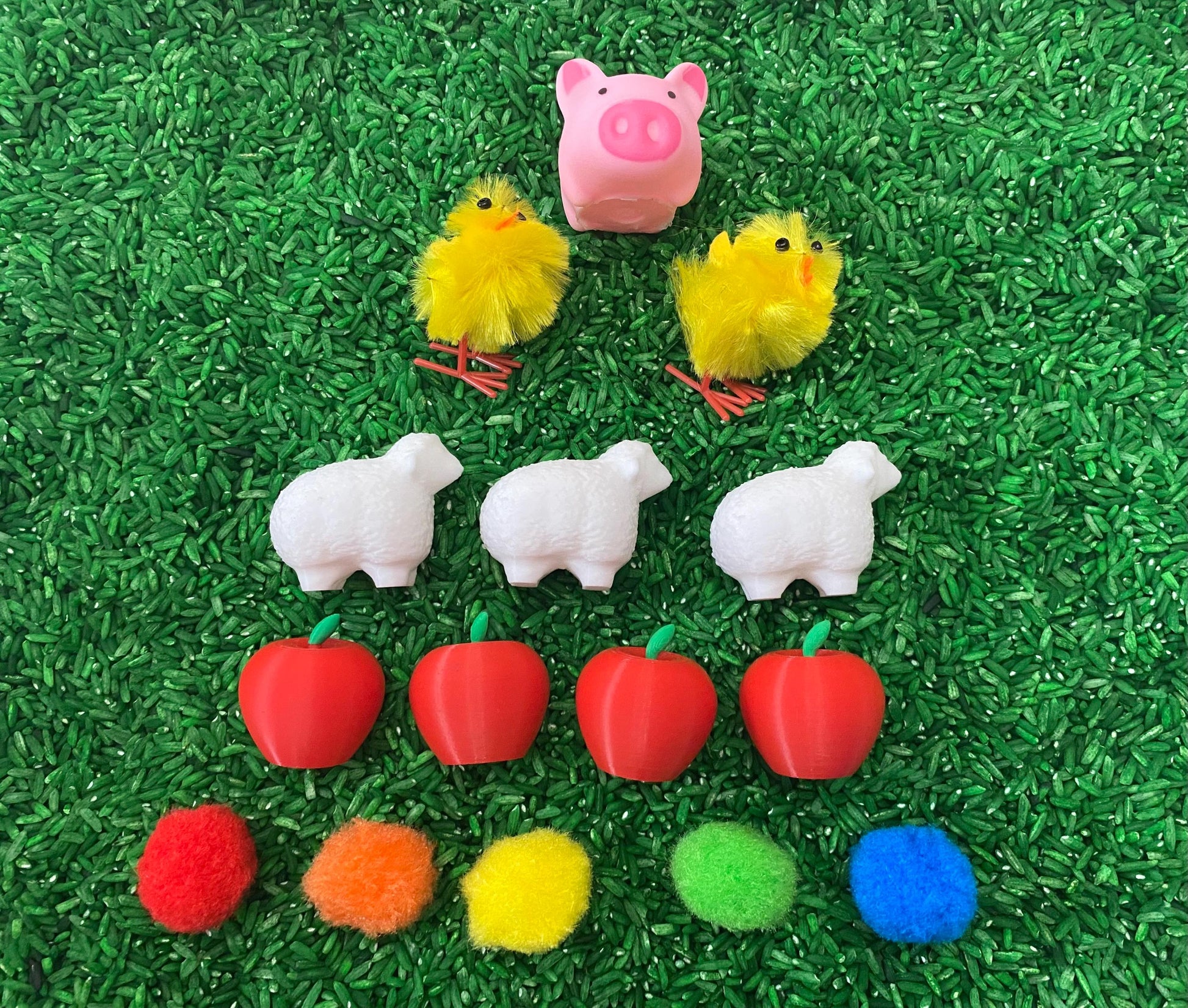 May include: A farm-themed sensory bin with a pink pig, yellow chicks, white sheep, red apples, and colorful pom-poms.