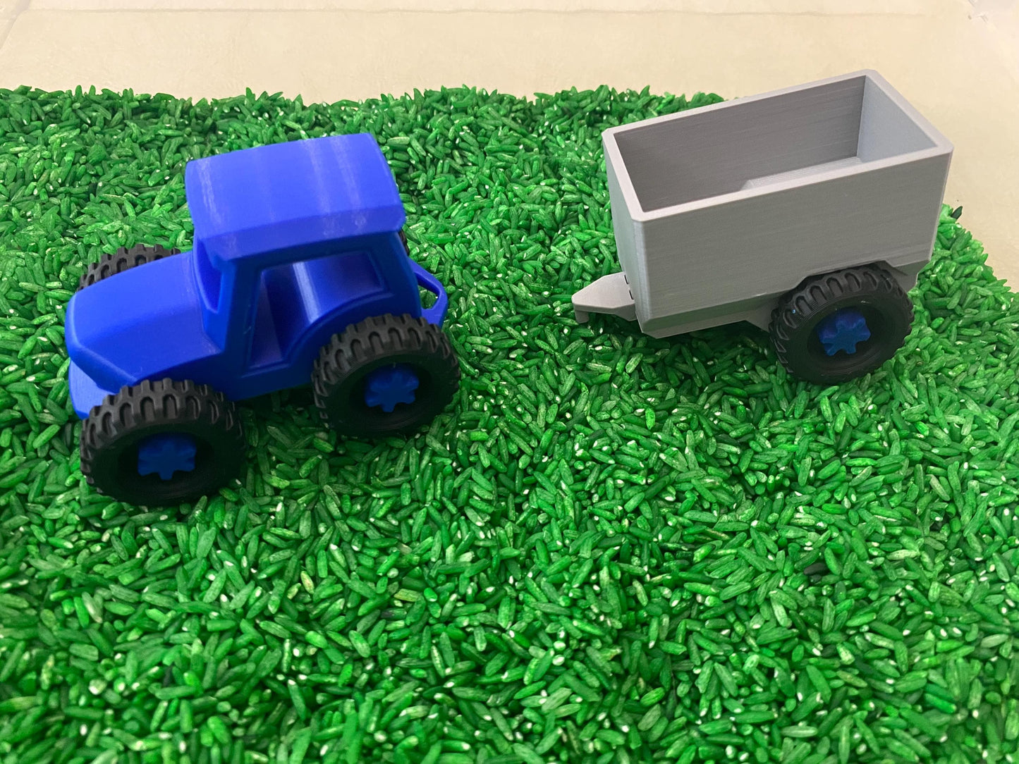 May include: A blue toy tractor with black wheels and a gray toy trailer with black wheels. The toys are on a bed of green rice.