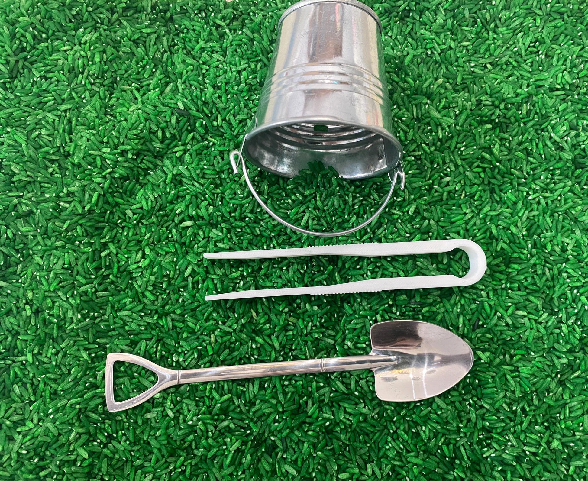 May include: A set of three silver-colored toy tools on a green background. The tools include a small bucket, a shovel, and a pair of tongs.