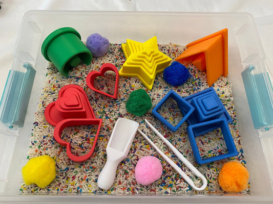May include: A clear plastic bin filled with sensory materials. Includes colorful shapes like hearts, stars, and squares, along with pom-poms, a scoop, and tongs. The base is filled with rice and colorful sprinkles. A fun, educational toy.