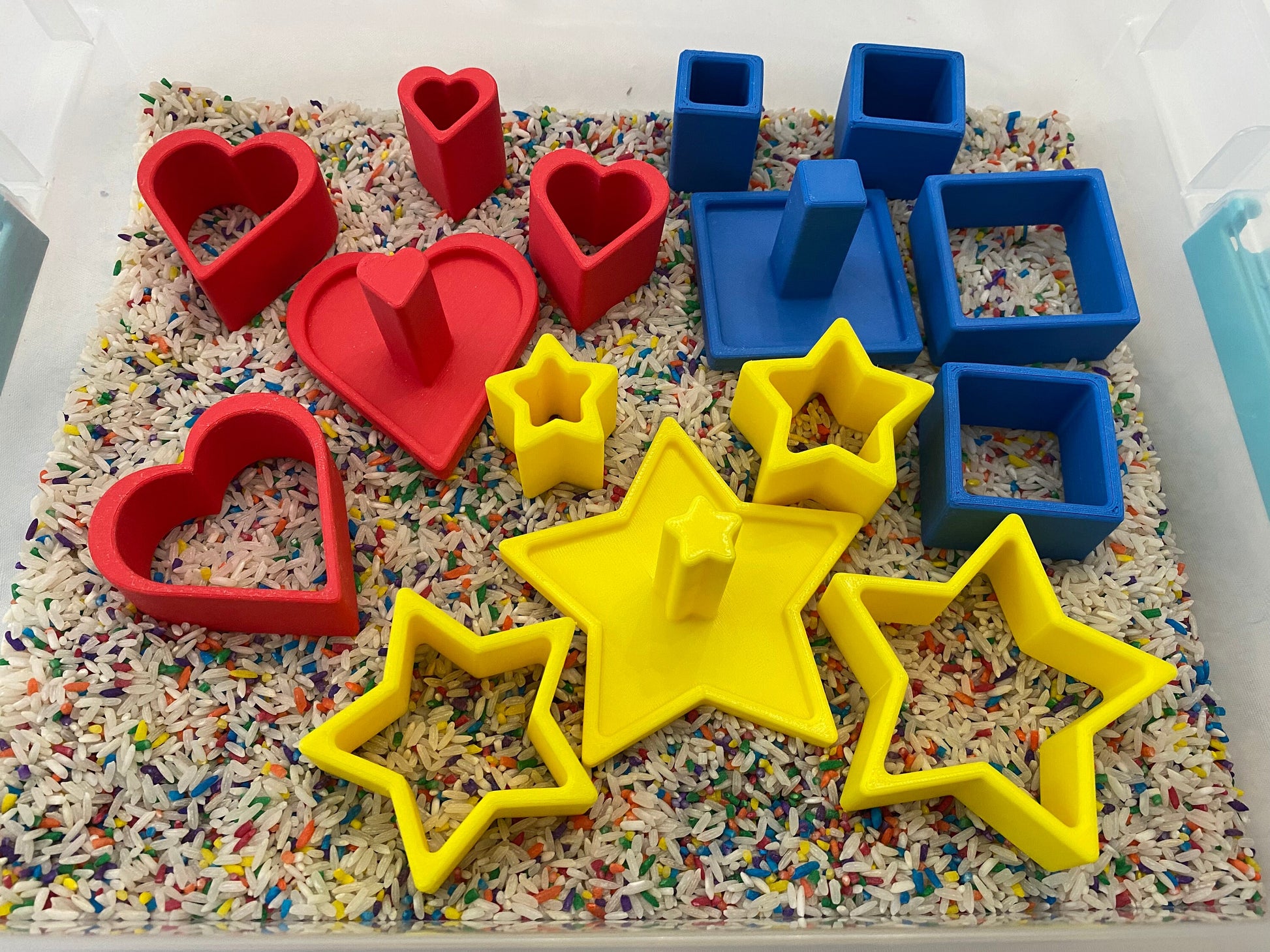 May include: A sensory bin filled with colorful rice and various shapes. Red heart and blue square shapes are mixed with yellow star shapes. The shapes are made of plastic and are designed for children's play and learning.