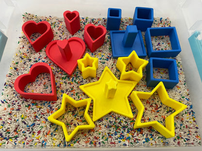 May include: A sensory bin filled with colorful rice and various shapes. Red heart and blue square shapes are mixed with yellow star shapes. The shapes are made of plastic and are designed for children's play and learning.