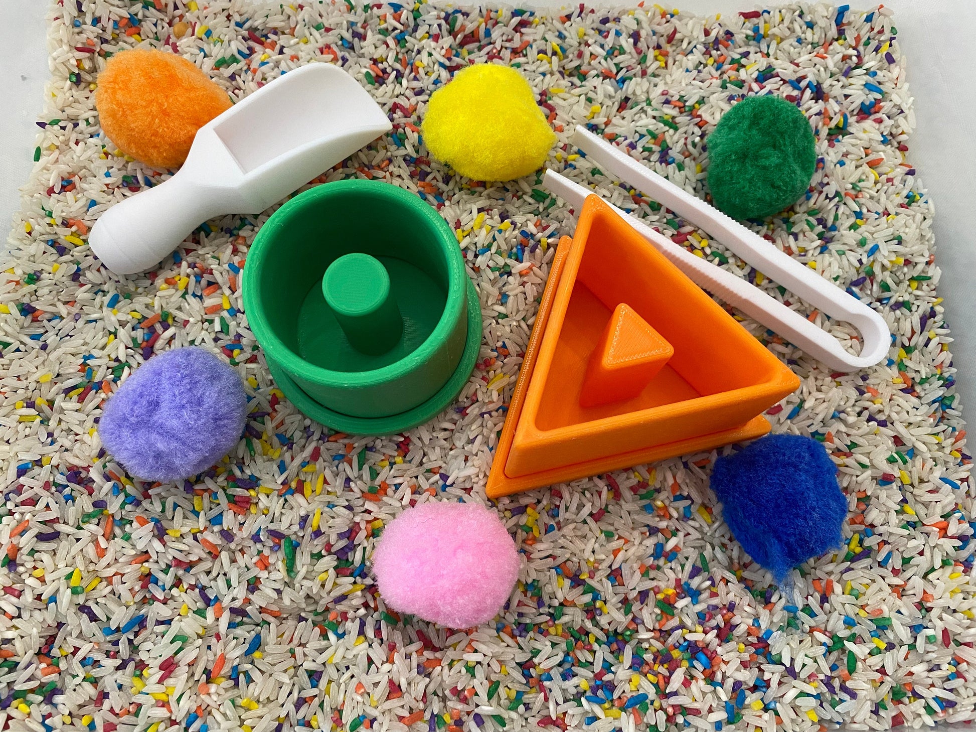May include: A sensory bin with various colorful elements. Includes a white scoop, green and orange geometric shapes, white tongs, and several fluffy pom-poms in orange, yellow, green, purple, and pink. The bin is filled with rice and colorful sprinkles.