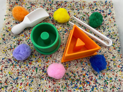 May include: A sensory bin with various colorful elements. Includes a white scoop, green and orange geometric shapes, white tongs, and several fluffy pom-poms in orange, yellow, green, purple, and pink. The bin is filled with rice and colorful sprinkles.