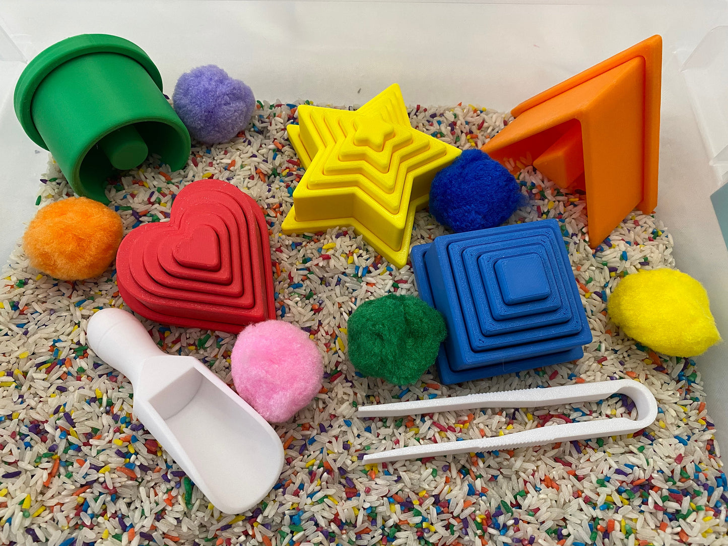 May include: A sensory bin filled with rice and colorful objects. Includes green, orange, yellow, red, and blue geometric shapes, pom-poms, a white scoop, and white tongs. The shapes include a heart, star, square, and triangle.