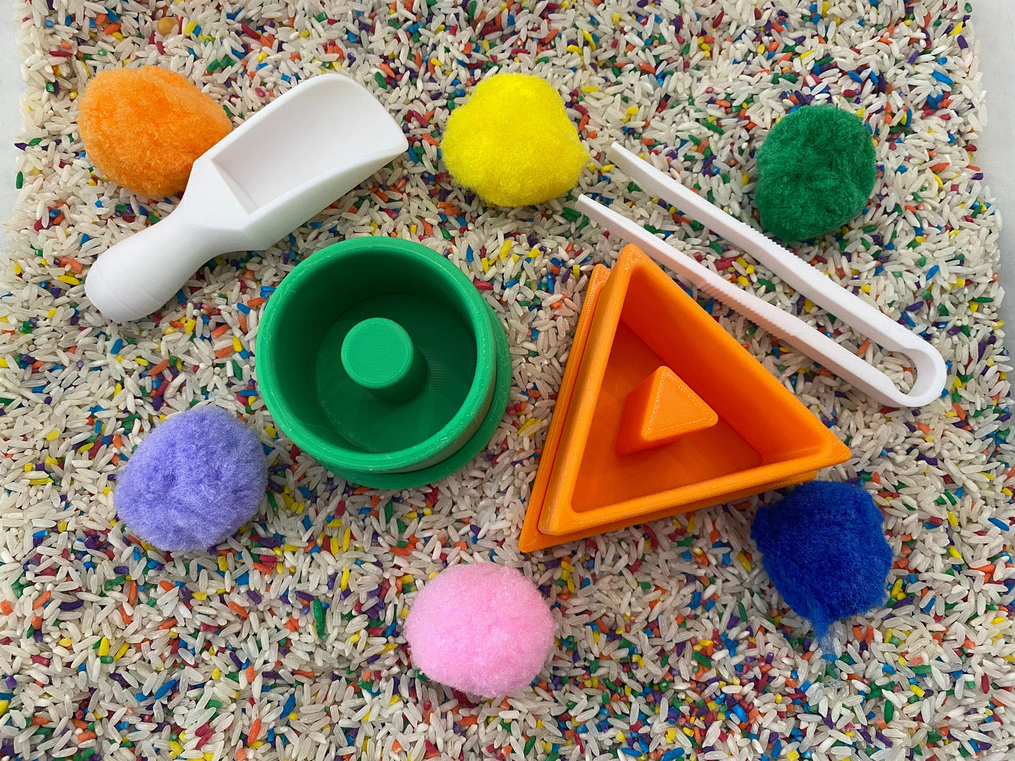 May include: A sensory bin with colorful pom-poms, a white scoop, green and orange geometric shapes, and white tongs. The bin is filled with rice speckled with colorful confetti. Educational toys for toddlers.