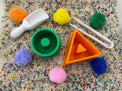 May include: A sensory bin with colorful pom-poms, a white scoop, green and orange geometric shapes, and white tongs. The bin is filled with rice speckled with colorful confetti. Educational toys for toddlers.
