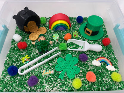 May include: A green sensory bin filled with white rice and green rice, with a variety of colorful pom-poms, a rainbow, a pot of gold coins, a leprechaun figurine, a shamrock, a green hat, and a white scoop.